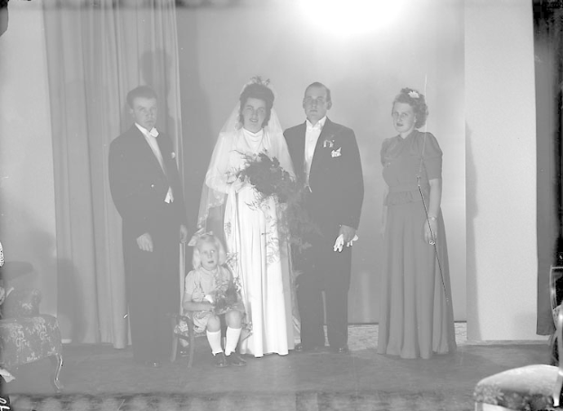 Ein Schwarz-Weiß-Foto einer Braut in einem weißen Kleid und Schleier, die einen Strauß hält, neben einem Bräutigam in einem schwarzen Anzug und weißem Hemd, mit zwei Erwachsenen und zwei Kindern um sie herum auf einer Bühne, mit einem Vorhang und einer Wand im Hintergrund.