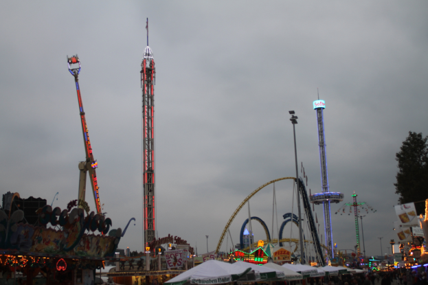 Eine Menschenmenge versammelt sich um einen hell erleuchteten Fasching in der Nacht, mit beleuchteten Fahrgeschäften, Pfählen und Bäumen unter einem bewölkten Himmel.