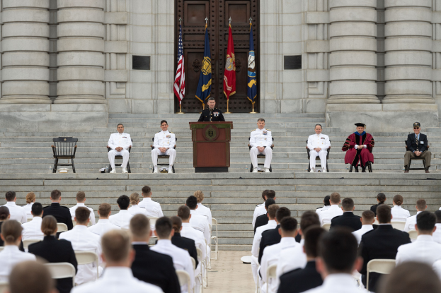 Gruppe von Menschen auf Stühlen vor einem Gebäude mit Säulen und einer Tür, mit einem Podium und Mikrofon in der Mitte und Flaggen im Hintergrund, was auf eine Abschlusszeremonie hinweist.