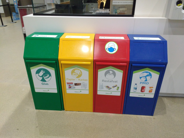 Eine Reihe von vier farbenfrohen Mülltonnen neben einem Tresen mit einem Monitor, anderen Gegenständen und einer Person links daneben.