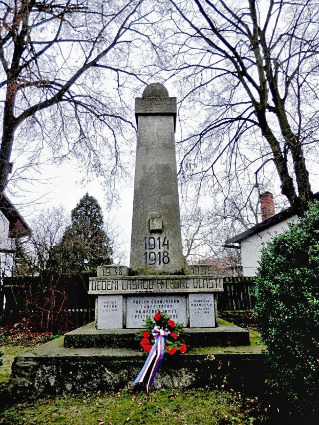 Ein Weltkriegsdenkmal auf einem Friedhof, geschmückt mit einem Kranz, umgeben von Gras, trockenen Blättern, Pflanzen und Bäumen, mit Häusern und Himmel im Hintergrund.