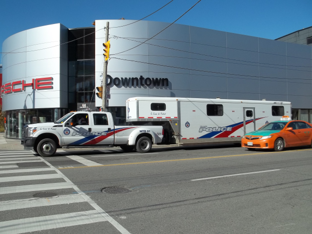 Ein Polizei-Lkw vor einem Gebäude mit Text geparkt, im Vordergrund ein Verkehrssignal mit einem Mast und Drähten und im Hintergrund Bäume mit einem klaren blauen Himmel.