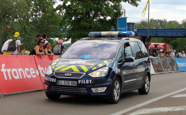 Ein Polizeiwagen fährt auf einer Straße neben einer Menschenmenge, einige tragen Kappen und tägliche Taschen, mit einem Banner links, Gelönd mit Bannern dahinter und einem Hintergrund mit Bäumen, einer Brücke, einer Fahne auf einem Mast und einem bewölktem Himmel.