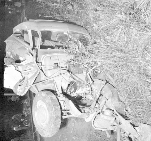 Schwarz-weißes Foto eines stark beschädigten Autos mit eingedellter Front und zersplitterten Fenstern, das auf der Seite einer Straße mit einem Baum und verstreuten Gegenständen im Hintergrund liegt.