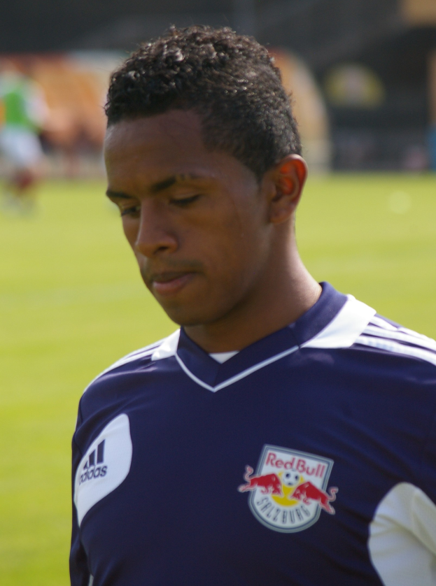 Ein junger Mann in einem blauen T-Shirt steht auf einem Fußballfeld und schaut entschlossen nach vorne, mit kurzem dunklem Haar.