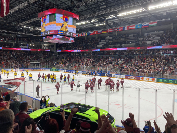 Ein Eishockeyspiel in einer großen Arena mit Zuschauern, ein Auto vor der Menge, Bande, Banner, Deckenbeleuchtung, ein Bildschirm und hängende Flaggen.