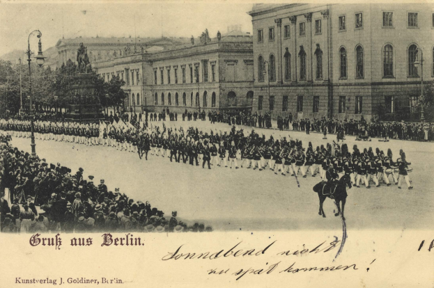 Schwarz-weiß-Foto von einem historischen Umzug in Berlin mit Menschen, Reiter zu Pferde, einer Statue, Straßenelementen, Gebäuden und einem bewölkten Himmel.