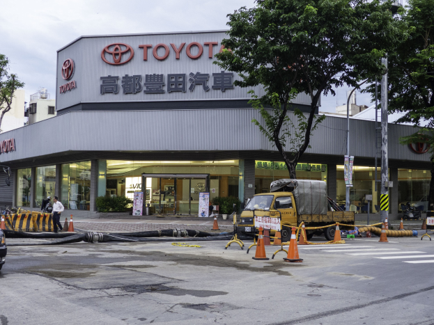 Ein Toyota-Autohaus in Tokio, Japan, mit Fahrzeugen, Verkehrskegeln, Rohren, Menschen, Bäumen, Polen, Brettern und Gebäuden im Vordergrund und einem bewölkten Himmel im Hintergrund.