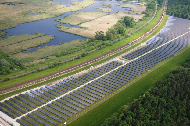 Luftbild einer Solaränlage mit Panelen, umgeben von Bäumen, Gras, Wasser und einer näheren Schiene.