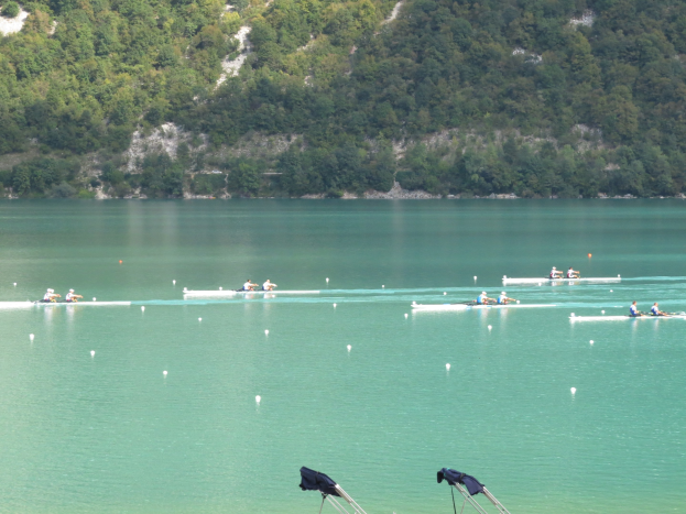 Gruppe von Menschen bei einem Ruderwettbewerb auf einem Gewässer mit Bäumen und einem Hügel im Hintergrund, Kleidung über Stühle am Boden gehängt.