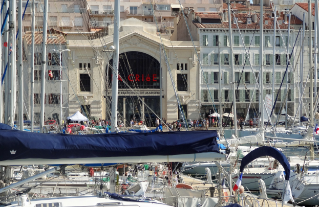 Ein Hafen mit zahlreichen Booten im Wasser, Gebäude im Hintergrund mit Fenstern und einem Namensschild sowie Zelte und Menschen.