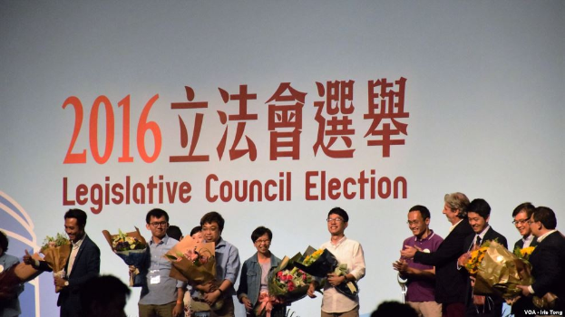 Eine Gruppe von Menschen auf einer Bühne mit Blumensträußen, umgeben von einer Zuschauermenge und einem Bildschirm mit Text und Logo im Hintergrund, der die Legislativratswahl 2016 anzeigt.