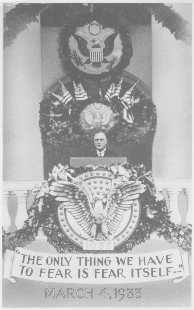 Schwarzes und weißes Foto eines Mannes, der an einem Rednerpult spricht, mit einem Kranz und einem Emblem an der Wand dahinter und der Inschrift "Das einzige, wovor wir uns fürchten müssen, ist die Furcht selbst" unten.