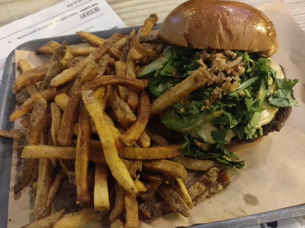 Ein Tablett mit einem Hamburger, der mit Salat, Tomate und Zwiebeln belegt und mit goldenen, knusprigen Pommes frites garniert ist, das auf einem Tisch mit einem Blatt Papier daneben steht.
