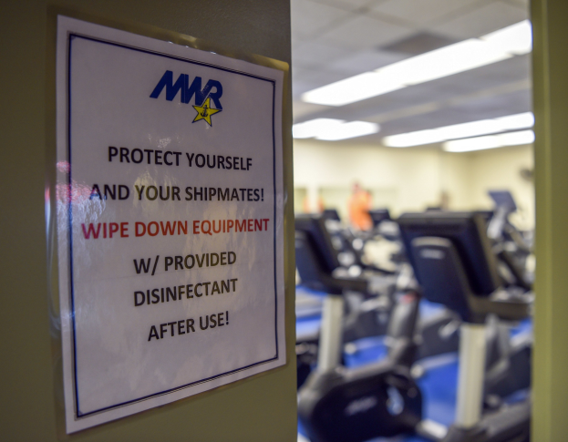 Ein Schild an einer Turnhalle mit der Aufschrift "Schützen Sie sich und Ihre Schiffskameraden" mit Laufbändern und Deckenlampen im Hintergrund, das Bild leicht unscharf, um Bewegung anzudeuten.