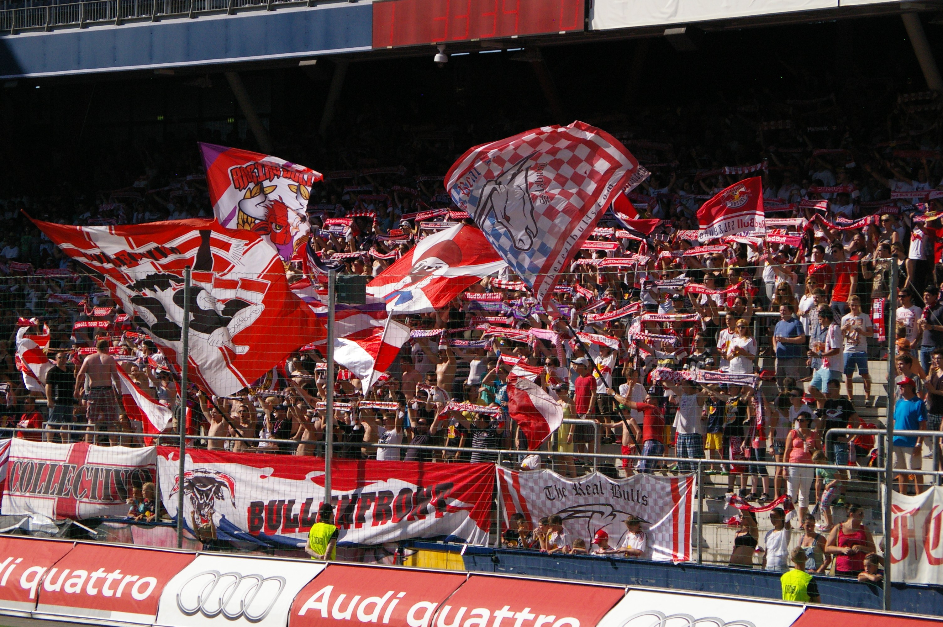 Eine große Menschenmenge in einem Stadion mit Fahnen, Bannern, einem Metallzaun, einer Anzeigetafel, einem Bildschirm und Deckenleuchten.