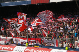 Eine große Menschenmenge in einem Stadion mit Fahnen, Bannern, einem Metallzaun, einer Anzeigetafel, einem Bildschirm und Deckenleuchten.