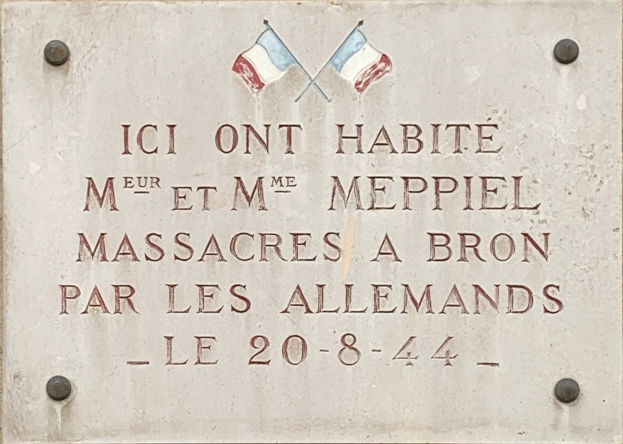 Steintafel an der Fassade eines Gebäudes mit zwei Flaggen darüber, mit Inschrift, die ein Massaker gedenkt.