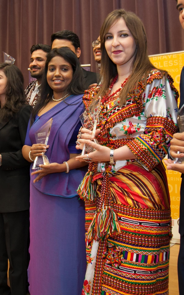 Eine Gruppe von Menschen auf einer Bühne mit einer Frau in der Mitte, die eine Trophäe hält, dahinter ein Vorhang und eine Tafel mit der Aufschrift "Bravo Segen Award Gewinner."