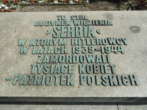 Gedenkstein mit eingravierter Plakette vor einem Gebäude umgeben von blühenden Pflanzen.