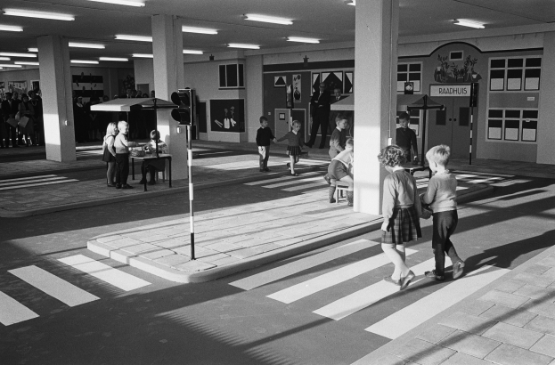 Schwarzes und weißes Foto von Kindern, die eine Straße an einer Ampel überqueren, mit Fußgängern in der Nähe, einem Verkehrssignalmast im Vordergrund und Säulen mit Deckenlampen im Hintergrund.