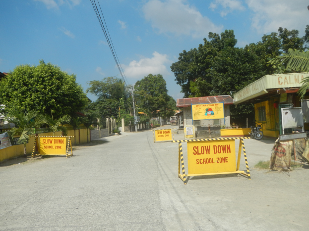 Eine Straße mit einem gelben "Slow Down School Zone"-Schild, geparkte Fahrzeuge auf der rechten Seite und ein Hintergrund mit Bäumen, Gebäuden, Polen, Drähten und dem Himmel.