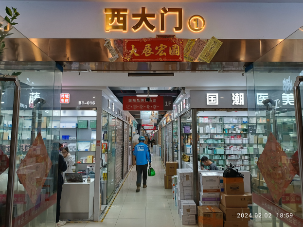 Innenraum einer chinesischen Apotheke mit Glaswänden, Regalen voller Artikel, verstreuten Kartons, einer Pflanze links und einer Namensschild oben.