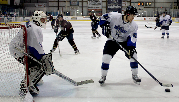 Eine Gruppe junger Männer, die auf einem Eisplatz Eishockey spielen, tragen Helme und halten Hockeystöcke in den Händen, links steht ein Torpfosten und im Hintergrund sind Banner zu sehen.