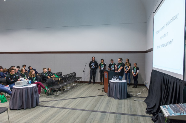 Eine Person, die in ein Mikrofon spricht, bei einem Treffen mit Teammitgliedern, die ID-Karten tragen, einem Präsentationsbildschirm und einem Videoplayer in einem leeren Büroraum.