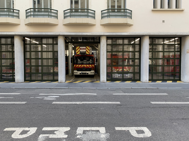 Eine Feuerwache mit einem Feuerwehrauto davor, mit Fenstern, Geländern, Säulen und Glastüren, mit einer Straße unten.