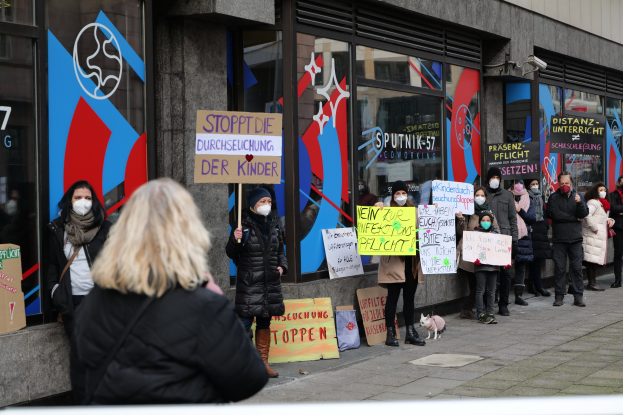 Eine Gruppe von Menschen mit Masken und Schildern protestiert vor einem Gebäude mit Glaswänden, begleitet von Texttafeln, Kameras und einem Hund.