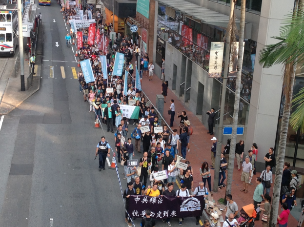 Eine große Gruppe von Menschen marschiert auf einer Straße in Hong Kong, hält Protestschilder und hat Bäume und glasummantelte Gebäude auf der rechten Seite und Fahrzeuge auf der linken Seite.