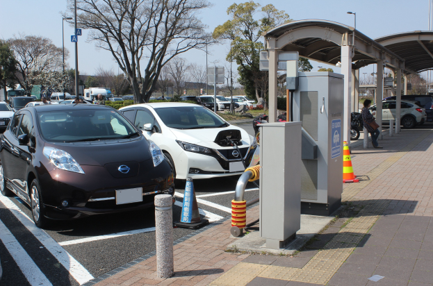 Ladestation für Elektroauto in Japan mit Autos auf der Straße, Verkehrskegel, einer Person auf dem Gehweg, einem Schuppen, Masten, Lichtern, Schildern, Bäumen, Pflanzen und einem Himmel im Hintergrund.