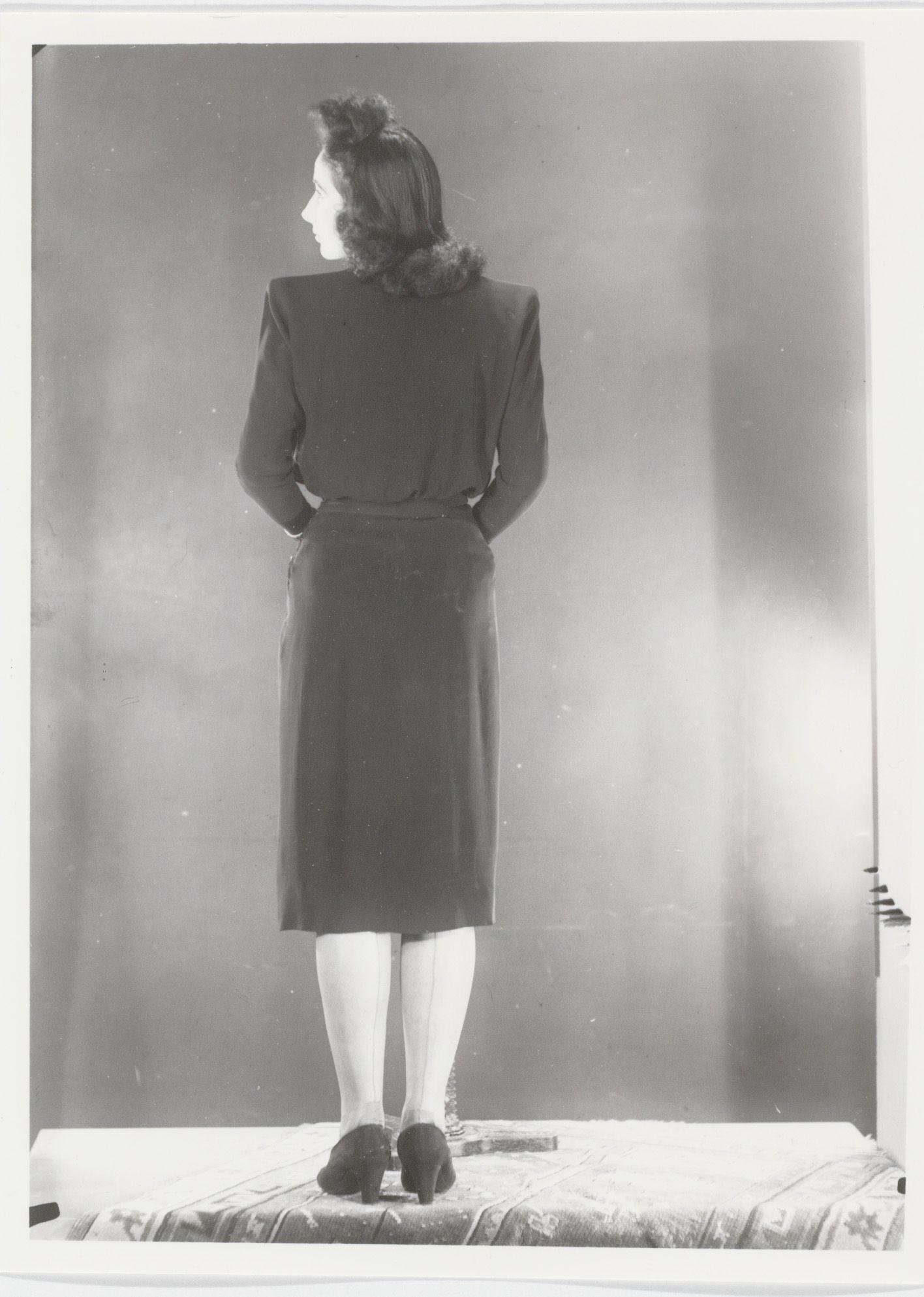 Ein Schwarz-Weiß-Foto einer Frau in einem Kleid im Stil der 1940er Jahre mit hohem Kragen, langen Ärmeln und Plisseerock, die schwarze High Heels trägt und eine Hochsteckfrisur hat, die auf einem Podest steht und zur Seite schaut mit einem leichten Lächeln.