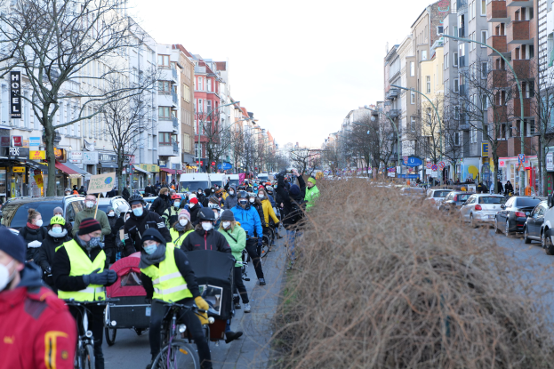 Eine große Gruppe von Menschen mit Masken und Sicherheitswesten auf Fahrrädern die eine straße mit Bäumen, Gebäuden, Lichtmasten und Texttafeln entlangfahren, neben Fahrzeugen und trockenem Gras unter einem klaren blauen Himmel.