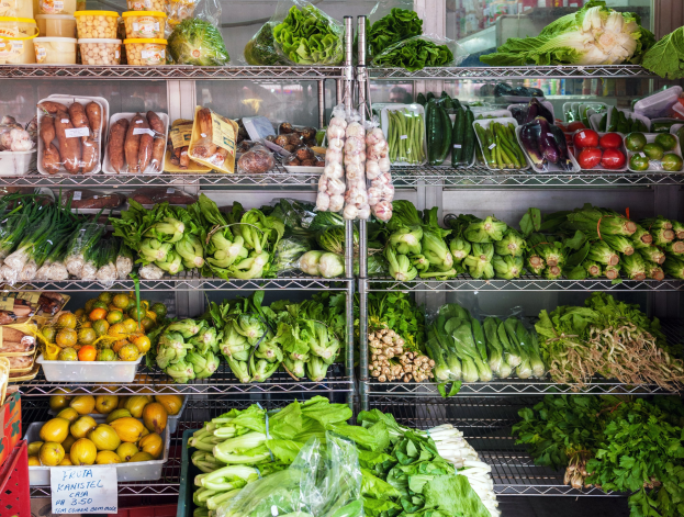 Ein Gang in einem Supermarkt mit frischem Gemüse und Obst unter Plastikabdeckungen, mit Kisten und einem Schild im Hintergrund und einem Glasfenster am Ende.