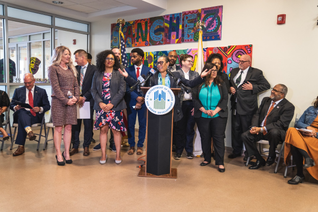 Eine Gruppe von Menschen steht vor einem Podium mit einem Mikrofon und einer Tafel mit der Aufschrift "Sancho-Stadtrat tritt dem Stadtrat bei", mit anderen im Hintergrund sitzend und stehend, Flaggen, einer Tafel, einem Feuerlöscher und Deckenleuchten an der Wand.
