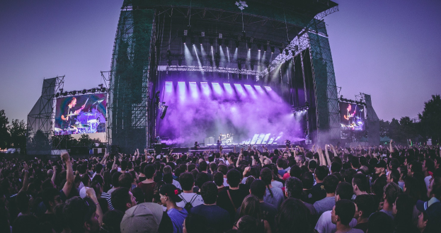 Eine große Menge Menschen auf einem Musikfestival schaut auf einer hell erleuchteten Bühne mit Instrumenten und Bildschirmen, die vor einem Gebäude, Bäumen und einem klaren blauen Himmel liegen.
