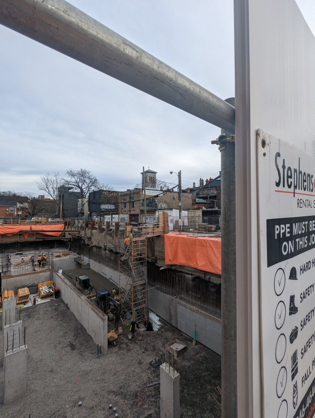 Baustelle mit einem Gebäude-Schild, das "Stephen's Rental Services" und eine Tafel auf der rechten Seite zeigt, vor Häusern, Bäumen, Pfählen und einem klaren blauen Himmel.