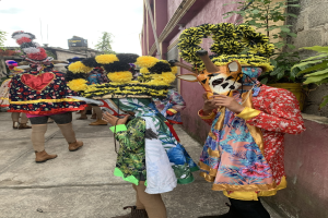 Eine Gruppe von Menschen in bunten Kostümen steht zusammen im Freien, mit verschiedenen Gegenständen, Bäumen, Gebäuden und einem klaren blauen Himmel im Hintergrund.