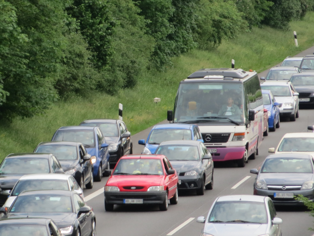 Verkehrsstau auf der Autobahn mit Autos und einem Lieferwagen, Menschen in Fahrzeugen, Bäume und Gras im Hintergrund.