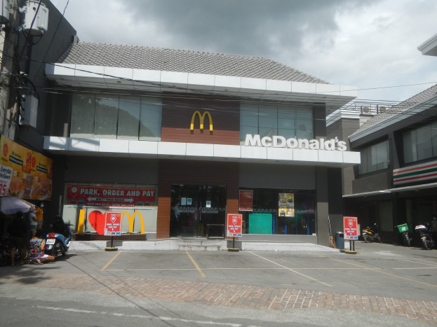 Ein McDonald's-Restaurant an einer Straßenecke mit umliegenden Gebäuden, Motorrädern, Fußgängern, Regenschirmen, Werbetafeln, Strommasten, Kabeln und einem bewölkten Himmel.