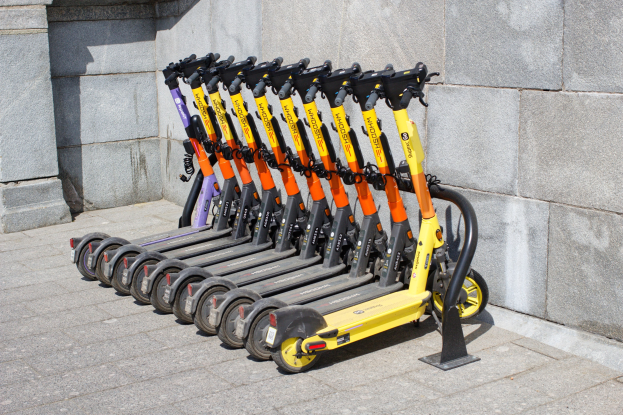 Eine Reihe von E-Scootern, die nebeneinander auf einem Gehweg geparkt sind, mit einer Wand im Hintergrund.