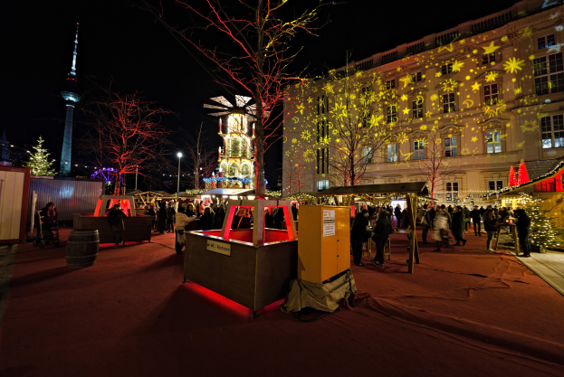 Ein geschäftiger Weihnachtsmarkt in Berlin, Deutschland, mit Menschen um geschmückte Stände, festliche Lichter, Bäume, Gebäude, Laternenpfähle und einen Turm unter einem dunklen Himmel.