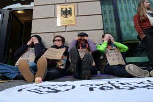 Eine Gruppe von Menschen sitzt vor einem Gebäude auf dem Boden, hält Schilder hoch, mit einem Banner und einem Apfel auf der Straße und einer Frau, die rechts steht und besorgt aussieht.