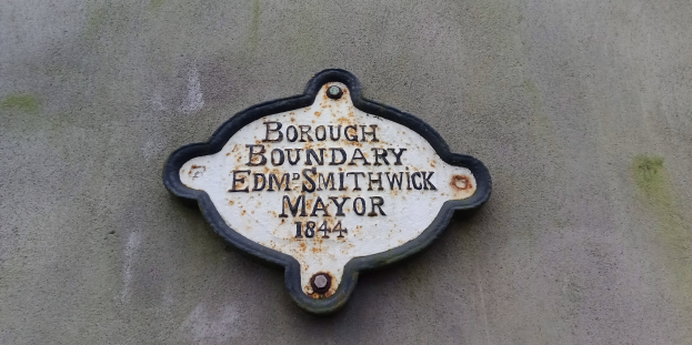 Metallschild an einer Gebäudeseite mit schwarzem Hintergrund und weißer fetter Schrift, die "Stadtgrenze Edm Smithwick Bürgermeister" lautet.