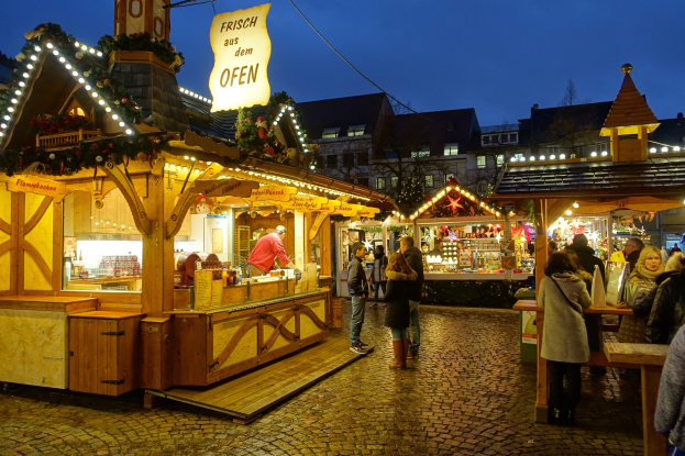 Ein belebter Weihnachtsmarkt auf Kopfsteinpflasterstraße bei Nacht mit Menschen um geschmückte Stände, Gebäude, Bäume und einen klaren Himmel im Hintergrund.