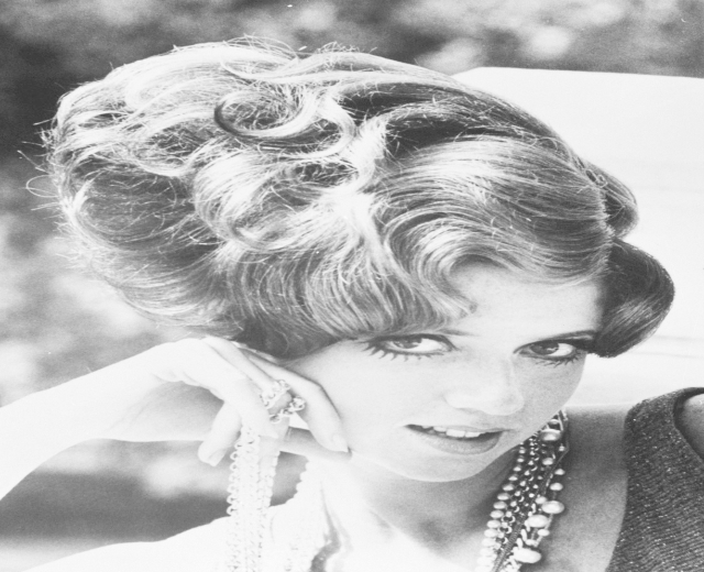 Schwarzes und weißes Foto von Prinzessin Diana in einem Kleid mit aufwendigen Accessoires, die vor einem unscharfen Hintergrund stehen.