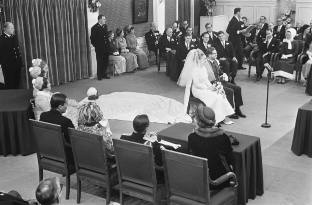Schwarz-weiß-Foto einer Hochzeitszeremonie mit sitzenden und stehenden Menschen, einem Mann mit Mikrofon, einer Frau mit Blumenstrauß, einem Podium mit Mikrofon rechts, Blumenschmuck und Vorhang links, einem gerahmten Foto und einer Tür im Hintergrund.