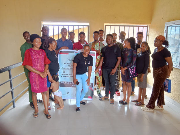 Eine Gruppe von Menschen, die nebeneinander in einem Raum stehen und eine Tafel in der Mitte halten, mit einem Geländer auf der linken Seite, Fenstern und einer Wand im Hintergrund.